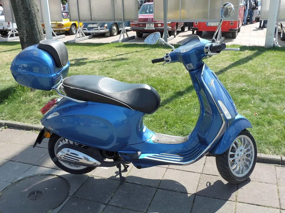 A blue Vespa scooter with a top box, parked on a pavement